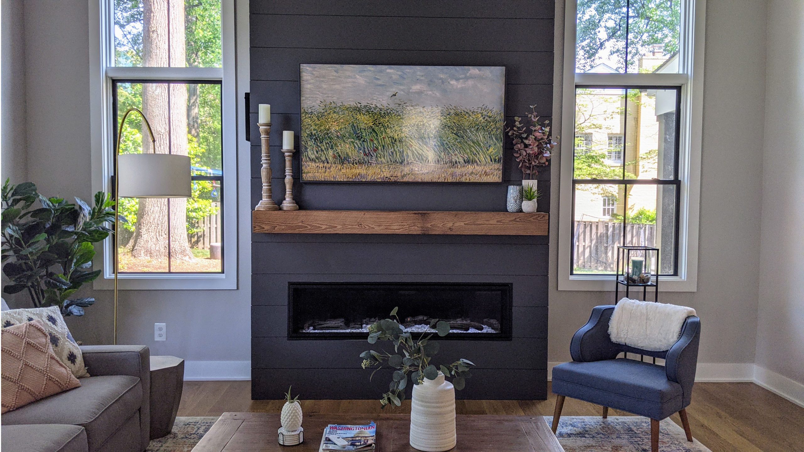 A brightly lit living room showing a fireplace with a reclaimed barn wood floating mantle above it. On the mantle are a couple vases and large decorative candle sticks. A TV floats above the mantle with an art piece on the screen.