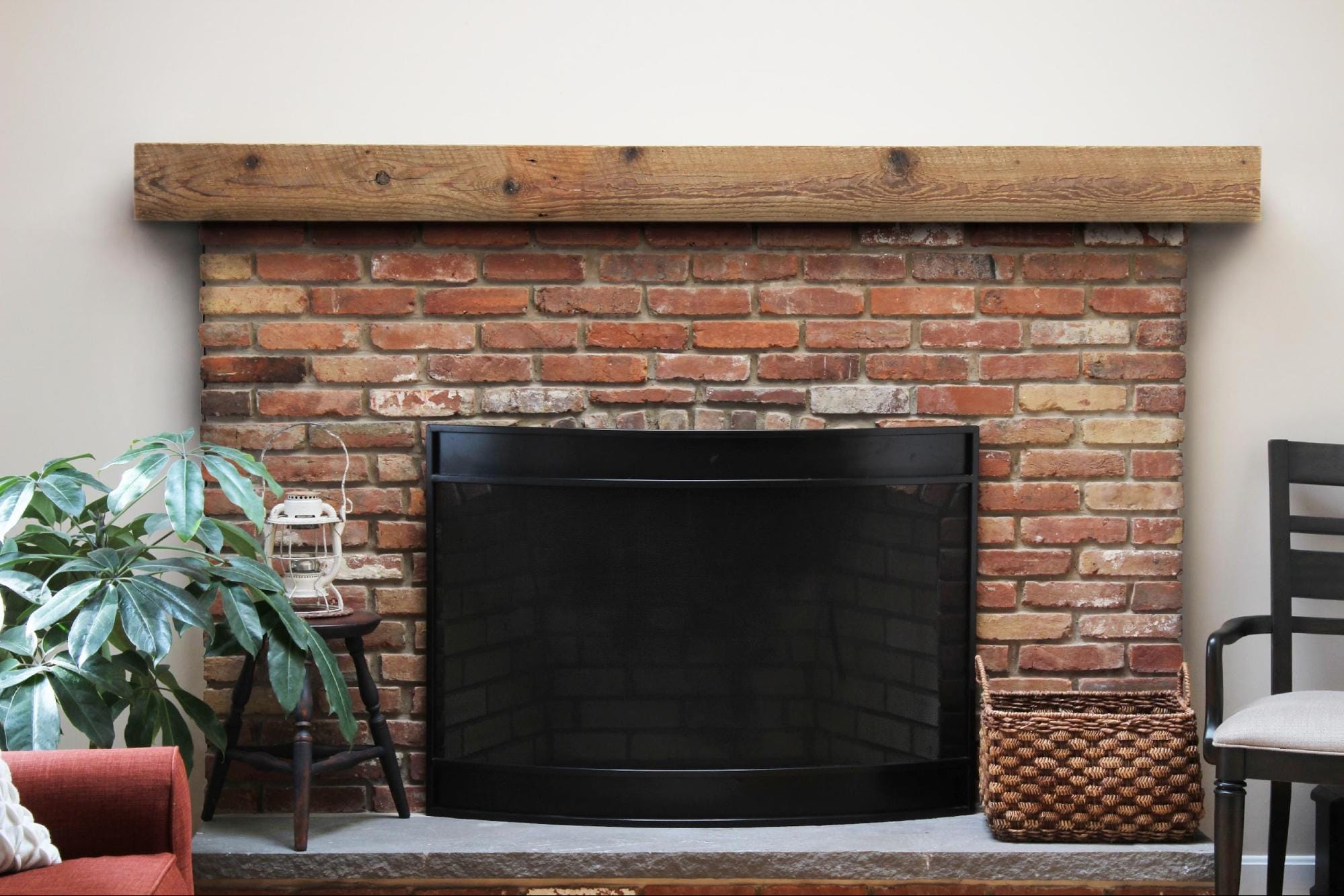 Rustic brick fireplace featuring a reclaimed wood box mantel with a black fireguard, wicker basket, and houseplant beside it.